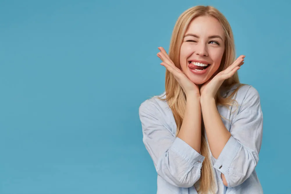 Mulher loira do lado direito da imagem com um fundo azul sorrindo enquanto coloca as mãos no rosto.